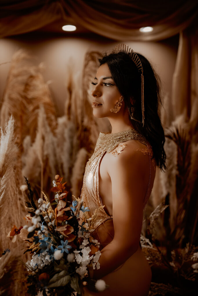 Woman covered in gold leaf in gold bodysuit holding bouquet of flowers for boudoir photoshoot