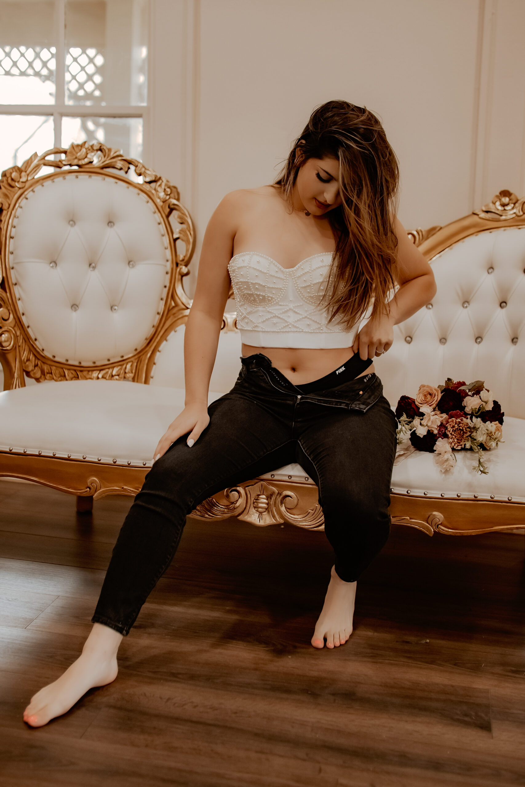 Woman with long brown hair looking down while sitting on a white vintage couch pulling on her black underwear