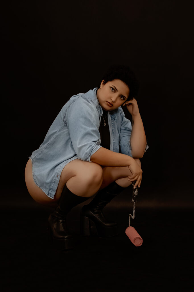 Boudoir outfit idea: client in oversized blue jean button-down shirt with black boots holding a paint brush  looking at camera